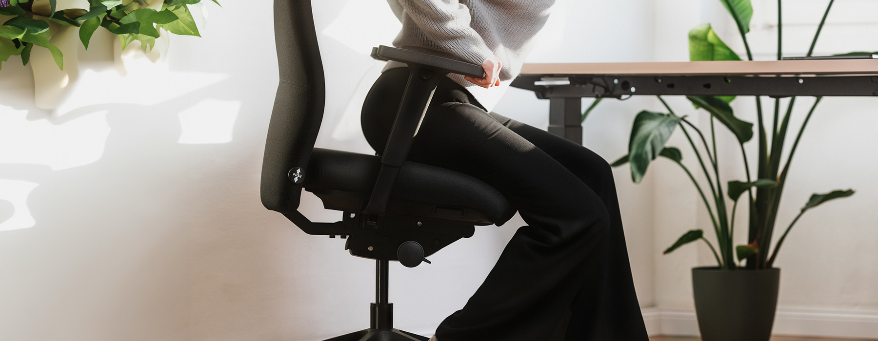 Person sitzt ergonomisch auf einem Bürostuhl in einem hellen Raum, mit einem höhenverstellbaren Schreibtisch im Hintergrund und Pflanzen als Dekoration.