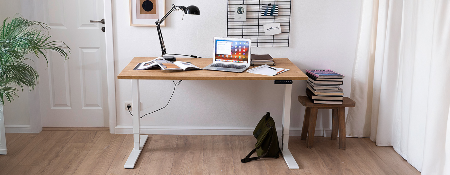 Moderner höhenverstellbarer Schreibtisch mit Holzplatte im Homeoffice, eingerichtet mit einem Laptop, Büchern, Notizheften und einer Schreibtischlampe. Im Hintergrund sind eine Zimmerpflanze und eine Pinnwand an der Wand zu sehen, was eine organisierte und produktive Arbeitsatmosphäre schafft.