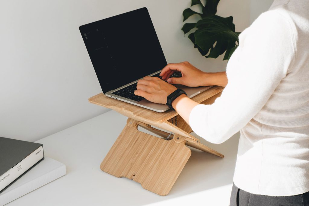 eine Person, die an einem Laptop arbeitet, der auf einem höhenverstellbaren Stehpultaufsatz Bambus platziert ist. Der Ständer hat ein minimalistisches Design mit klaren Linien und natürlicher Bambusmaserung.