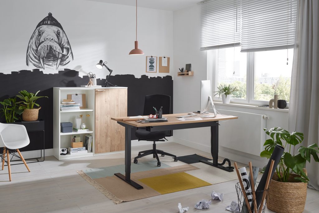 Modern eingerichtetes Homeoffice mit einem höhenverstellbaren Schreibtisch, großem Monitor, ergonomischem Stuhl und stilvollen Dekoelementen wie Pflanzen und Wandkunst. Der helle Raum hat ein großes Fenster, das viel Tageslicht hereinscheint, und schafft eine produktive und kreative Arbeitsatmosphäre.