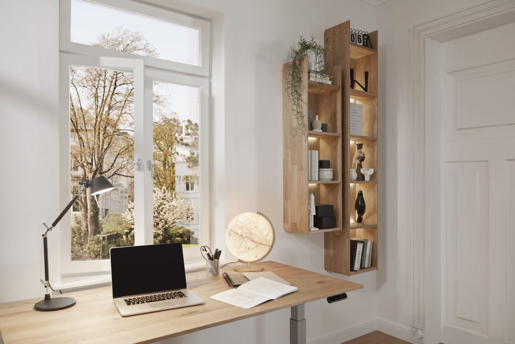 Ergonomischer Arbeitsplatz mit einem elektrisch höhenverstellbaren Schreibtisch, Laptop, Schreibtischlampe und dekorativen Regalen in einem modernen Homeoffice mit großem Fenster und Blick ins Grüne