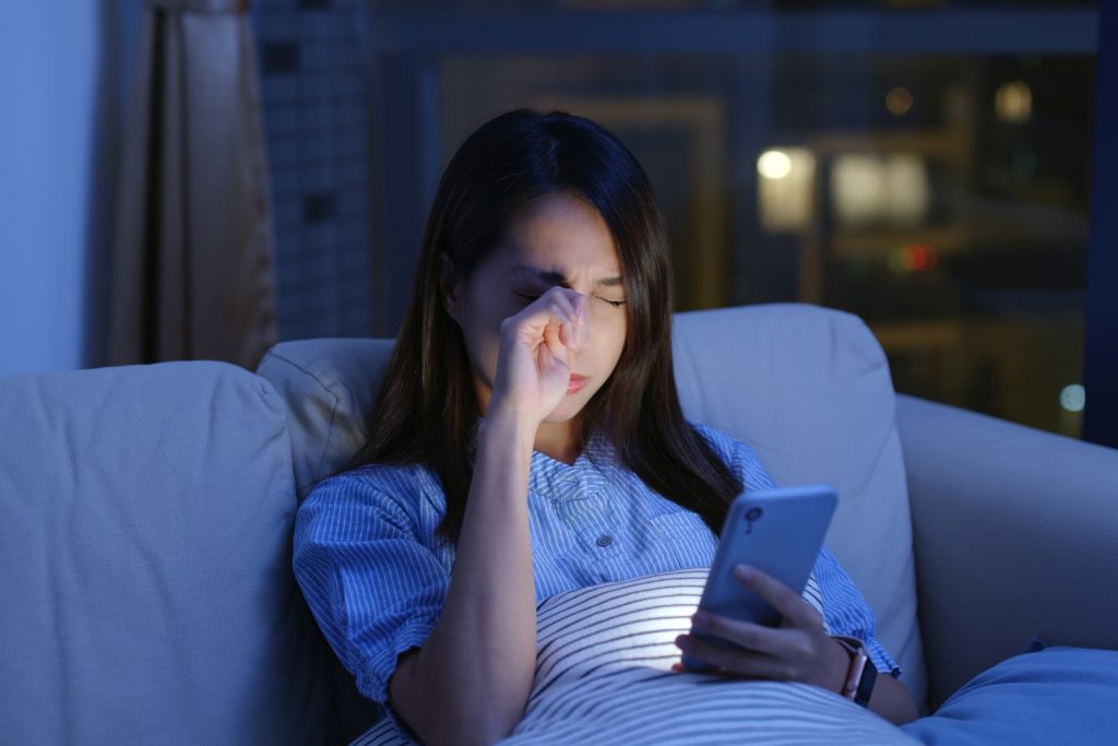 Person, die nachts auf einem Bett liegt und ein Smartphone in der Hand hält. Sie reibt sich müde die Augen, während das Licht des Bildschirms ihr Gesicht beleuchtet.