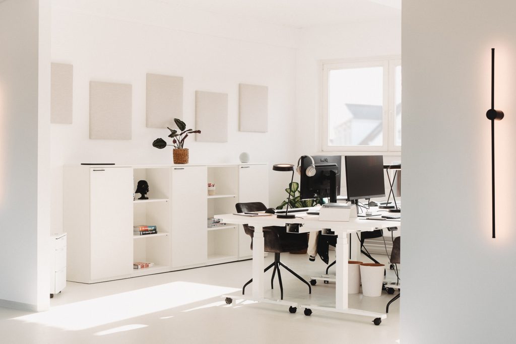 Helles, minimalistisches Büro mit höhenverstellbarem Schreibtisch auf Rollen und weißen Möbeln.